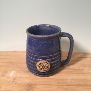 May include: A blue ceramic mug with a four-leaf clover design. The mug has a textured, striped design and a handle.