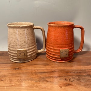 May include: Two ceramic mugs with a speckled glaze. One mug is beige with brown speckles and the other is orange with brown speckles. Both mugs have a raised design of the state of Washington on the front.