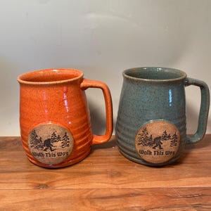 May include: Two ceramic mugs with a speckled glaze. The mugs are orange and blue-green. Each mug has a round, brown sticker with a cartoon Bigfoot and the text "Walk This Way".