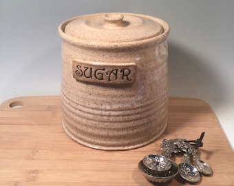 May include: A beige ceramic sugar canister with a lid and the word "SUGAR" on a rectangular plaque. The canister is on a wooden cutting board with a set of decorative measuring spoons.