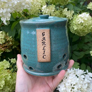 May include: A turquoise ceramic garlic keeper with a lid and small air holes. The word "GARLIC" is vertically printed on a rectangular label attached to the front. The keeper is held in front of a background of white and green flowers.