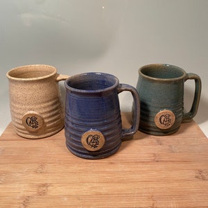 May include: Three ceramic mugs with a speckled glaze and a small monkey design on the front. The mugs are in shades of beige, blue, and green.