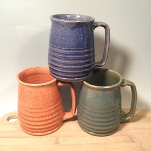 May include: Three ceramic mugs with different colored glazes. The mugs are stacked on top of each other. The top mug is blue, the middle mug is orange, and the bottom mug is green.