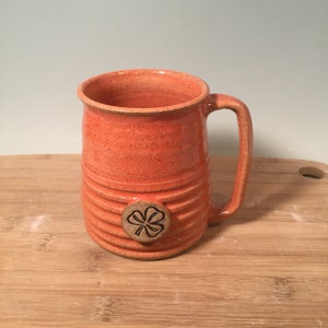 May include: A large orange ceramic mug with a brown four-leaf clover design. The mug has a textured, speckled finish and a wide, rounded handle.