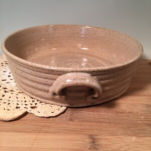 May include: A speckled, beige ceramic bowl with two curved handles. The bowl has a round shape with a slightly textured surface and a series of horizontal ridges around the exterior. The bowl is sitting on a wooden surface.