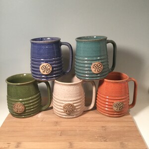 May include: Five ceramic mugs with a shamrock design. The mugs are in different colors: green, blue, white, orange, and teal.