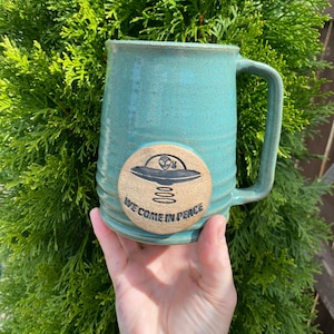 May include: A teal blue ceramic mug with a handle. The mug has a round, brown, ceramic sticker with a black and white illustration of a flying saucer and the text "We come in peace".