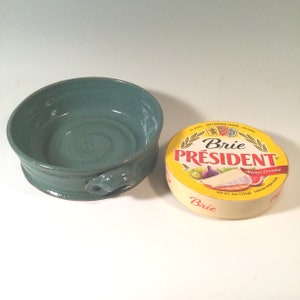 May include: A teal ceramic brie cheese baker with a handle next to a package of President Brie cheese. The cheese package is round with a yellow label and the word "Brie" in red.