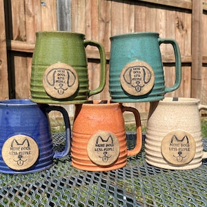 May include: Five ceramic mugs with a speckled glaze in various colors, including green, blue, orange, and beige. Each mug has a round, brown, ceramic tag with the text "More Dogs Less People" and a dog face graphic.