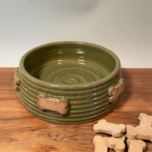 May include: A green ceramic dog bowl with a spiral design in the center. The bowl has a ribbed texture and is decorated with bone-shaped accents. Several dog biscuits are scattered nearby on a wooden surface.
