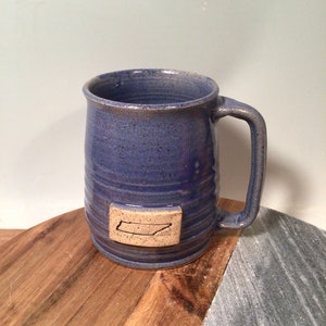 May include: A blue ceramic mug with a brown Tennessee state outline on the front. The mug is sitting on a wooden surface.