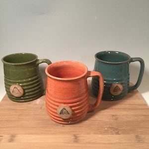 May include: Three ceramic mugs with a Celtic knot design. The mugs are green, orange, and blue.