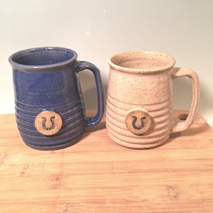May include: Two ceramic mugs with a horseshoe design. One mug is blue with white speckles and the other is white with brown speckles.