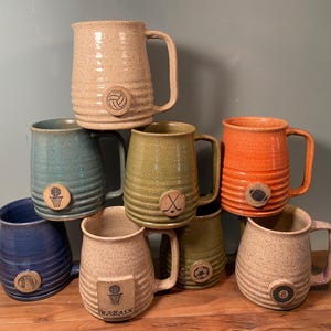 May include: A collection of handmade ceramic mugs in various colors, including tan, blue, green, and orange. Each mug features a sports-themed emblem, such as a basketball, volleyball, or hockey stick. The mugs are stacked on a wooden surface.