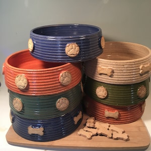 May include: A stack of six ceramic pet bowls in various colors, including blue, orange, green, and beige. The bowls have paw print and bone designs. A pile of dog biscuits is in the foreground.