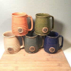 May include: Set of five ceramic mugs with a tree logo. The mugs are in various colors, including orange, green, beige, blue, and gray.