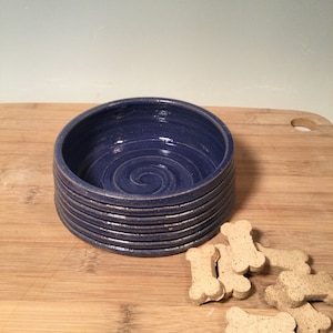 May include: A handmade, blue ceramic dog bowl with a spiral design in the center. The bowl has a textured, ribbed exterior and is accompanied by several bone-shaped dog treats. The bowl sits on a wooden cutting board.