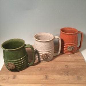 May include: Three ceramic mugs with a shamrock design. The mugs are green, white, and orange. The mugs are on a wooden surface.