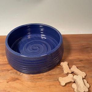 May include: A deep blue ceramic dog bowl with a spiral design in the center. The bowl has a wide, round shape with horizontal ridges. Several bone-shaped dog treats are next to the bowl on a wooden surface.
