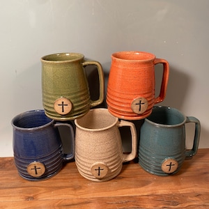 May include: Five ceramic mugs with a cross design on the front. The mugs are in different colors: green, orange, white, blue, and teal.