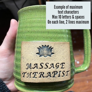 May include: Green ceramic mug with a brown rectangular label. The label has a black lotus flower design and the text "MASSAGE THERAPIST".