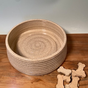 May include: A beige ceramic dog bowl with a speckled finish and a ridged exterior. Several bone-shaped dog treats are scattered nearby. The bowl is designed for pet food or water.