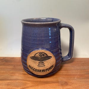 May include: A blue ceramic mug with a brown clay disc on the front. The disc has a black and white illustration of a flying saucer with the text "We Come In Peace".