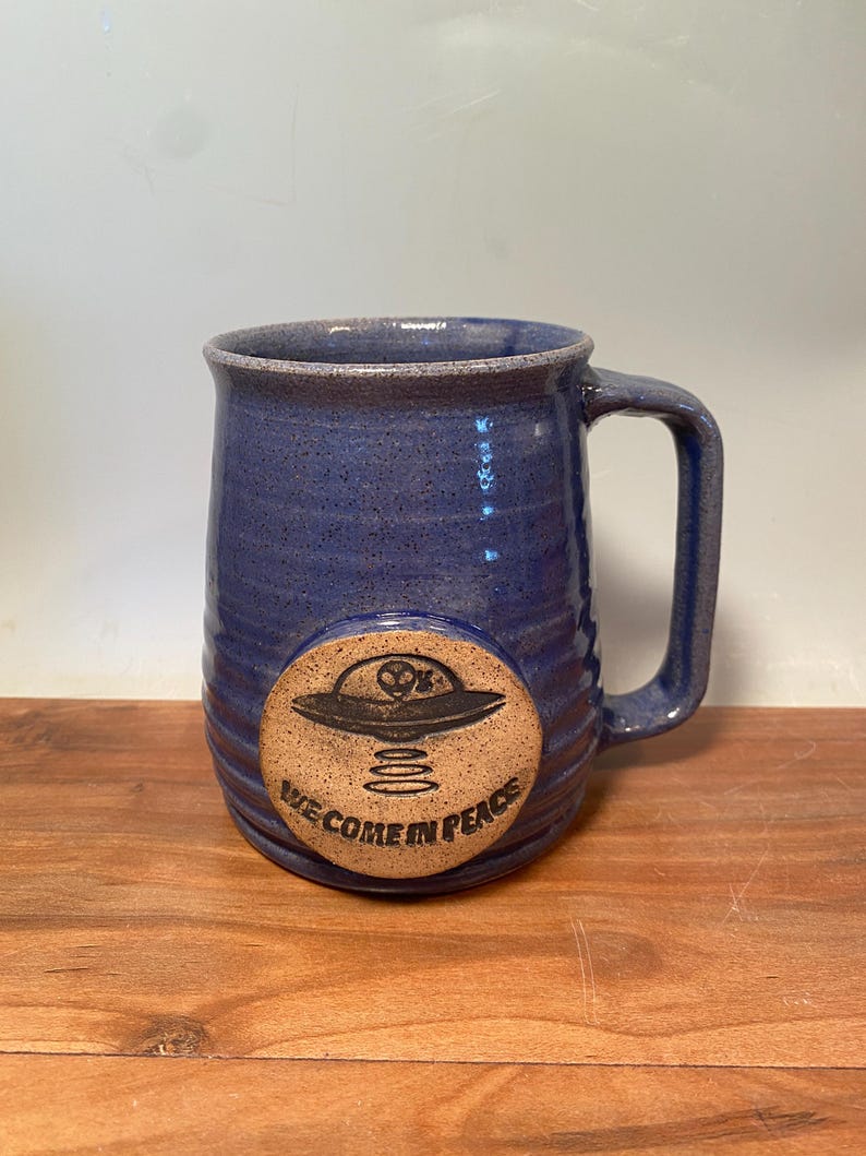 May include: A blue ceramic mug with a brown clay disc on the front. The disc has a black and white illustration of a flying saucer with the text "We Come In Peace".