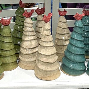 May include: A collection of ceramic Christmas tree figurines in various colors, including green, beige, and teal. Each tree has a spiral design and is topped with a red bird. The trees are arranged on a white shelf.