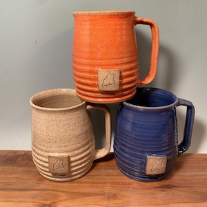 May include: Three ceramic mugs with a speckled glaze. The mugs are stacked on top of each other. The top mug is orange, the middle mug is beige, and the bottom mug is blue. Each mug has a small square with the outline of a state on it.