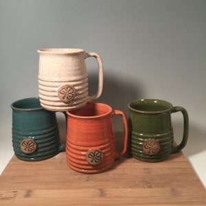 May include: Four ceramic mugs with a four-leaf clover design. The mugs are in white, green, orange, and brown.