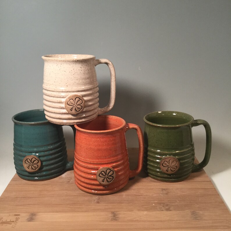 May include: Four ceramic mugs with a four-leaf clover design. The mugs are in white, green, orange, and brown.