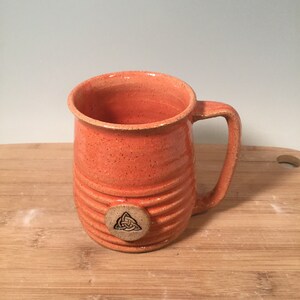 May include: A handmade orange ceramic mug with a textured, ribbed design. The mug has a handle and a circular design with a Celtic knot on the front.