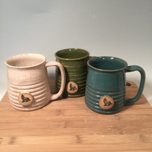 May include: Three ceramic mugs with a speckled glaze and a wolf design. The mugs are in white, green, and blue.