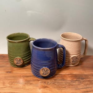 May include: Three ceramic mugs with a speckled glaze. The mugs are green, blue, and beige. Each mug has a small, round, brown emblem with a bird design on the front.