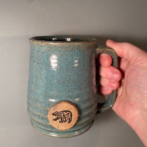 May include: A blue ceramic mug with a brown bear design on the side. The mug has a speckled glaze and a rounded handle.