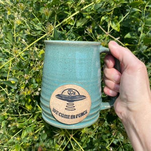 May include: A teal green ceramic mug with a brown circular sticker featuring a black alien spaceship and the text "We Come In Peace".