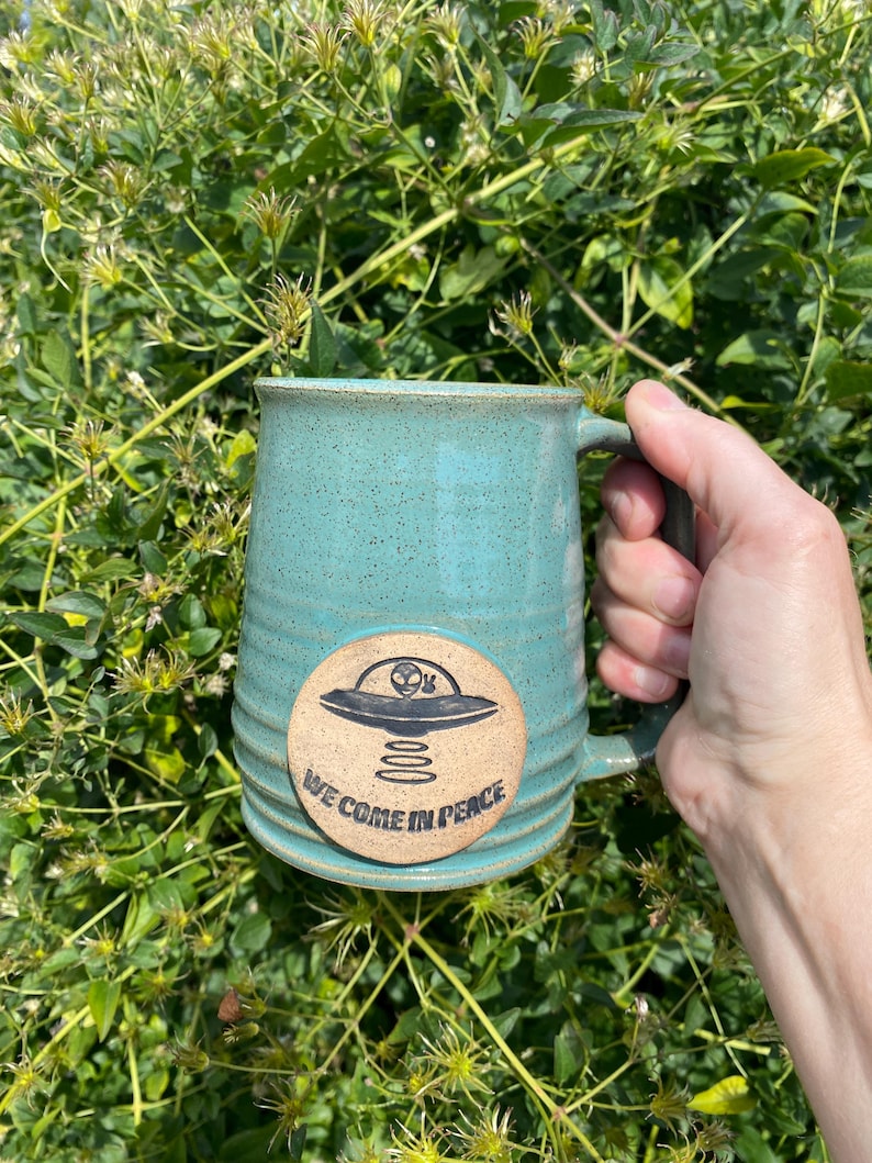 May include: A teal green ceramic mug with a brown circular sticker featuring a black alien spaceship and the text "We Come In Peace".