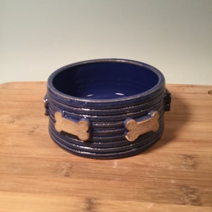 May include: A blue ceramic dog bowl with bone-shaped details around the rim. The bowl is on a wooden surface.