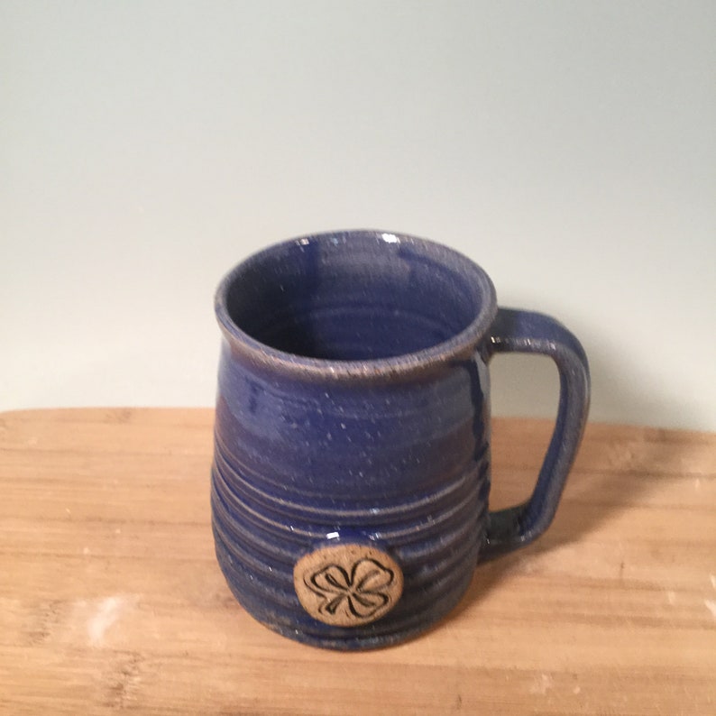 May include: A blue ceramic mug with a four-leaf clover design. The mug has a rounded body and a handle.