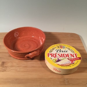 May include: An orange ceramic bowl with a handle sits next to a package of Brie cheese. The cheese package is round with a yellow label and the text "Brie President" and "Always Creamy". The cheese weighs 8 oz (226g).