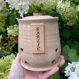 May include: A beige ceramic garlic keeper with a lid and holes for ventilation. The keeper has a wooden label that says "GARLIC" in black lettering.
