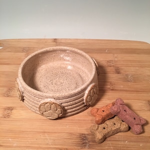 May include: A ceramic dog bowl with a speckled glaze and paw print designs around the rim. The bowl is sitting on a wooden surface with three dog biscuits.