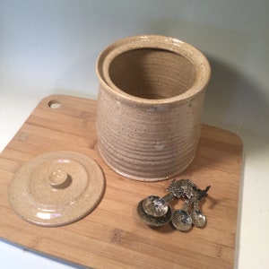 May include: A beige ceramic jar with a lid and a speckled glaze. The jar is sitting on a wooden cutting board with a set of four silver measuring spoons.