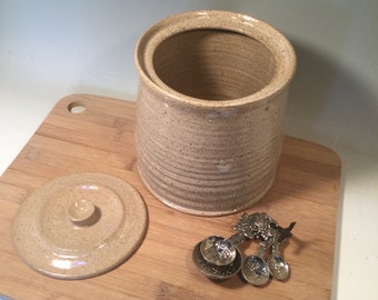 May include: A beige ceramic jar with a lid and a speckled glaze. The jar is sitting on a wooden cutting board with a set of four silver measuring spoons.