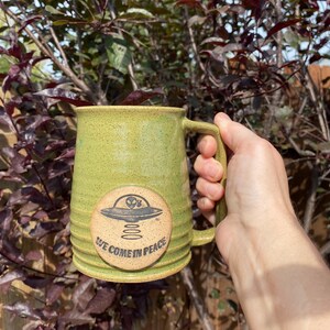 May include: Green ceramic mug with a brown handle. The mug has a circular design with a black alien spaceship and the text "We Come In Peace".