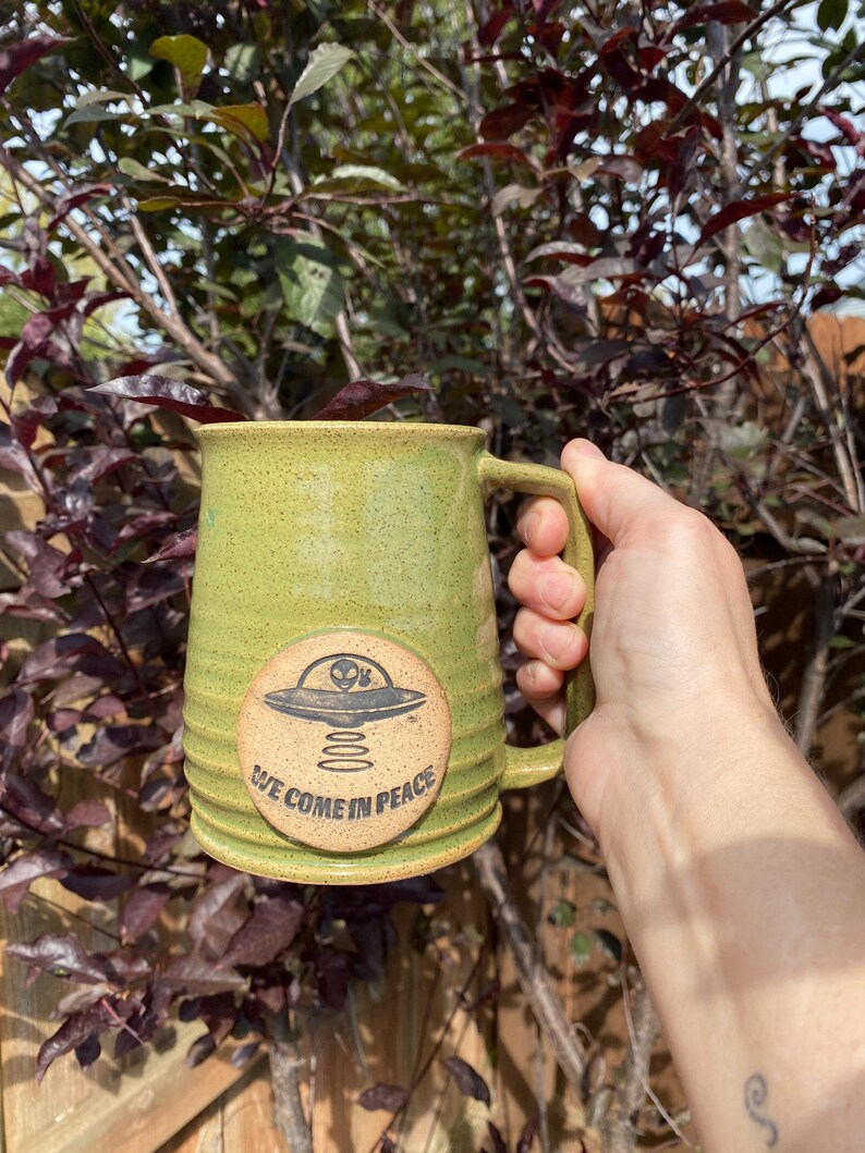 May include: Green ceramic mug with a brown handle. The mug has a circular design with a black alien spaceship and the text "We Come In Peace".