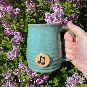 May include: A turquoise ceramic mug with a speckled finish and a handle. A wooden, circular emblem with a black musical note is affixed to the mug. The mug is held in front of a background of purple flowers and green leaves.