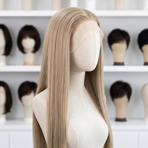 May include: A long, light blonde wig with a lace front is displayed on a mannequin head. The wig has a straight, sleek style and falls to the mid-chest. Other wigs are visible on shelves in the background.