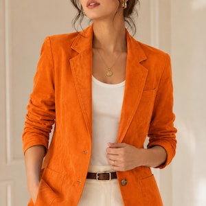 May include: A vibrant orange blazer with a textured finish, featuring a notched lapel, two front buttons, and two patch pockets. The blazer is worn over a plain white tank top and paired with white trousers secured by a brown leather belt.
